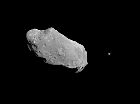 An image of asteroid Ida, a large, irregularly shaped space rock with visible craters, accompanied by its small moon Dactyl, both set against the blackness of space