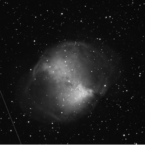 Black-and-white image of a nebula taken through a green filter, showing a round, cloud-like structure with areas of varying brightness. The nebula is set against a dense field of stars, with a faint diagonal line visible, possibly from a satellite or other object passing through the frame