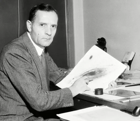 A black and white photograph of Edwin Hubble. He is sat at his desk, facing the camera, holding an image of a galaxy in his hands.