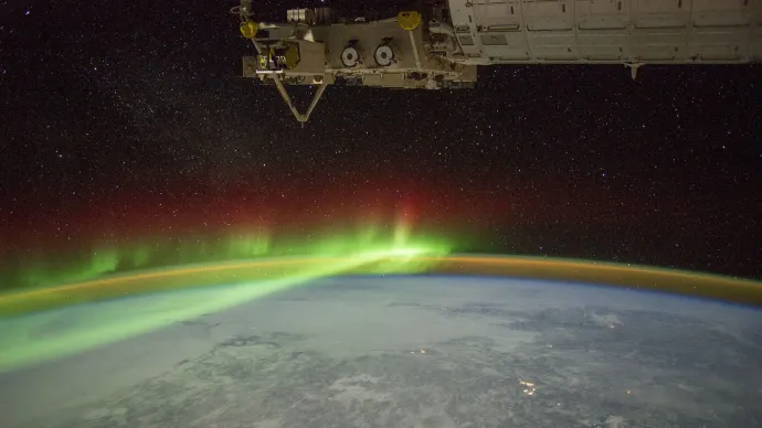 A view of earth from the international space station, part of which can be seen in the upper right of the image. A section of Earth can seen against a black background with stars visible. A green band of light is visible across the top of the Earth. 