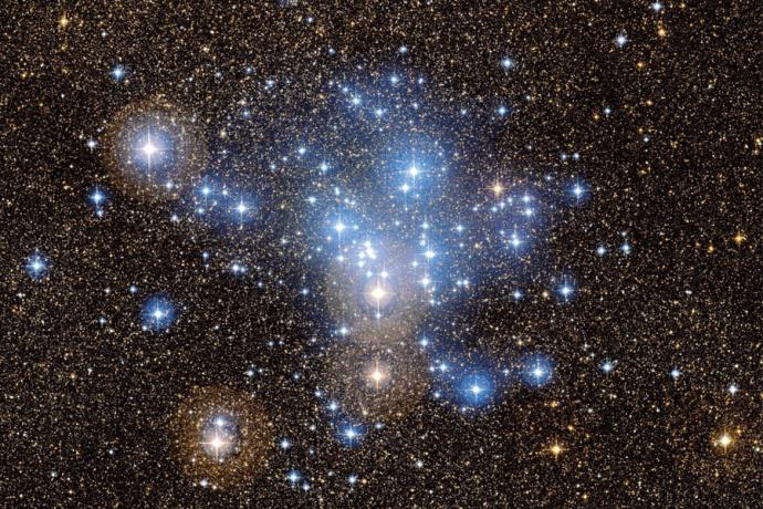 A photo of an open cluster of stars, several bright stars can be seen, mainly blue in colour, with some light being reflected from gas and dust close to the stars.