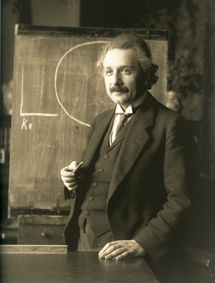 A sepia-toned photograph of Albert Einstein standing in front of a chalkboard, holding a piece of chalk. He is dressed in a formal suit and is slightly smiling, with part of a chalk drawing visible behind him