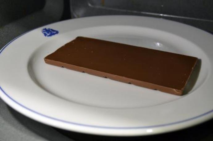 An unwrapped rectangular piece of chocolate placed on a white plate inside a microwave, ready for an experiment